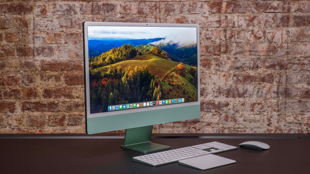 Apple iMac M3 in green front facing to the right with its color-matched accessories in front of it and sitting on a black desk in front of a brick wall.