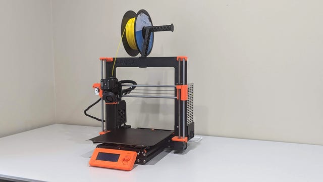 A black 3D printer with orange corners and a roll of yellow filament on top