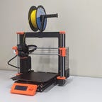 A black 3D printer with orange corners and a roll of yellow filament on top